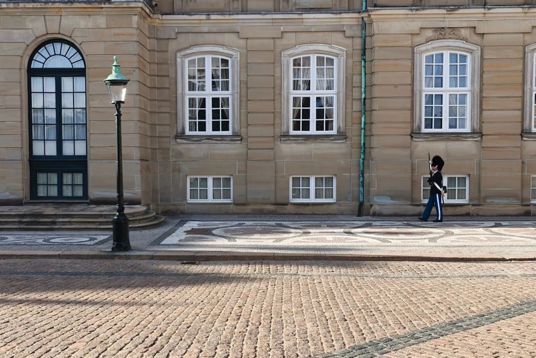 Stone building facade with arched and white windows, a lamppost, and a person walking on the sidewalk.