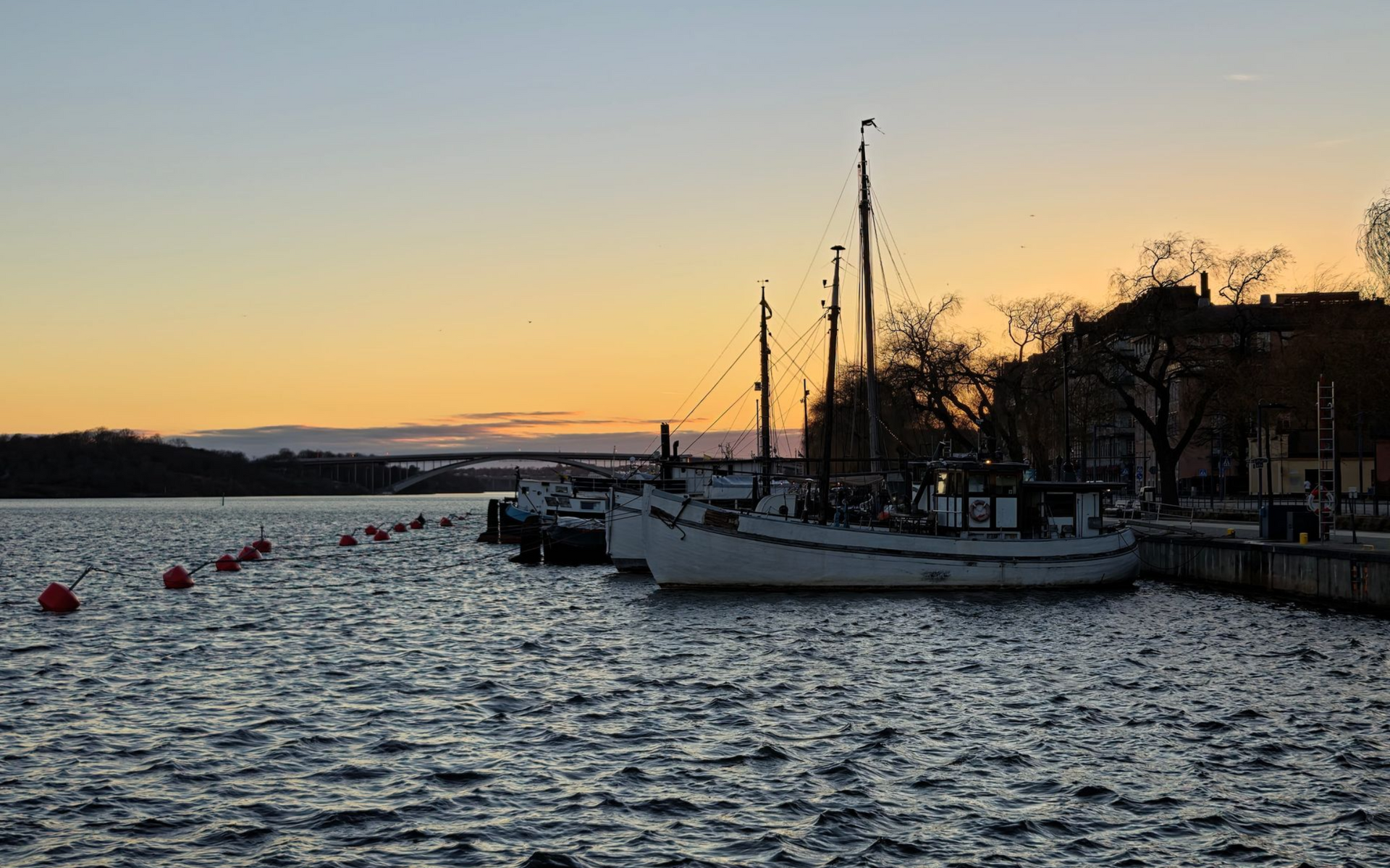 Stockholm Aussicht Sailboat at dusk on choppy water with orange sunset sky and city shoreline in silhouette