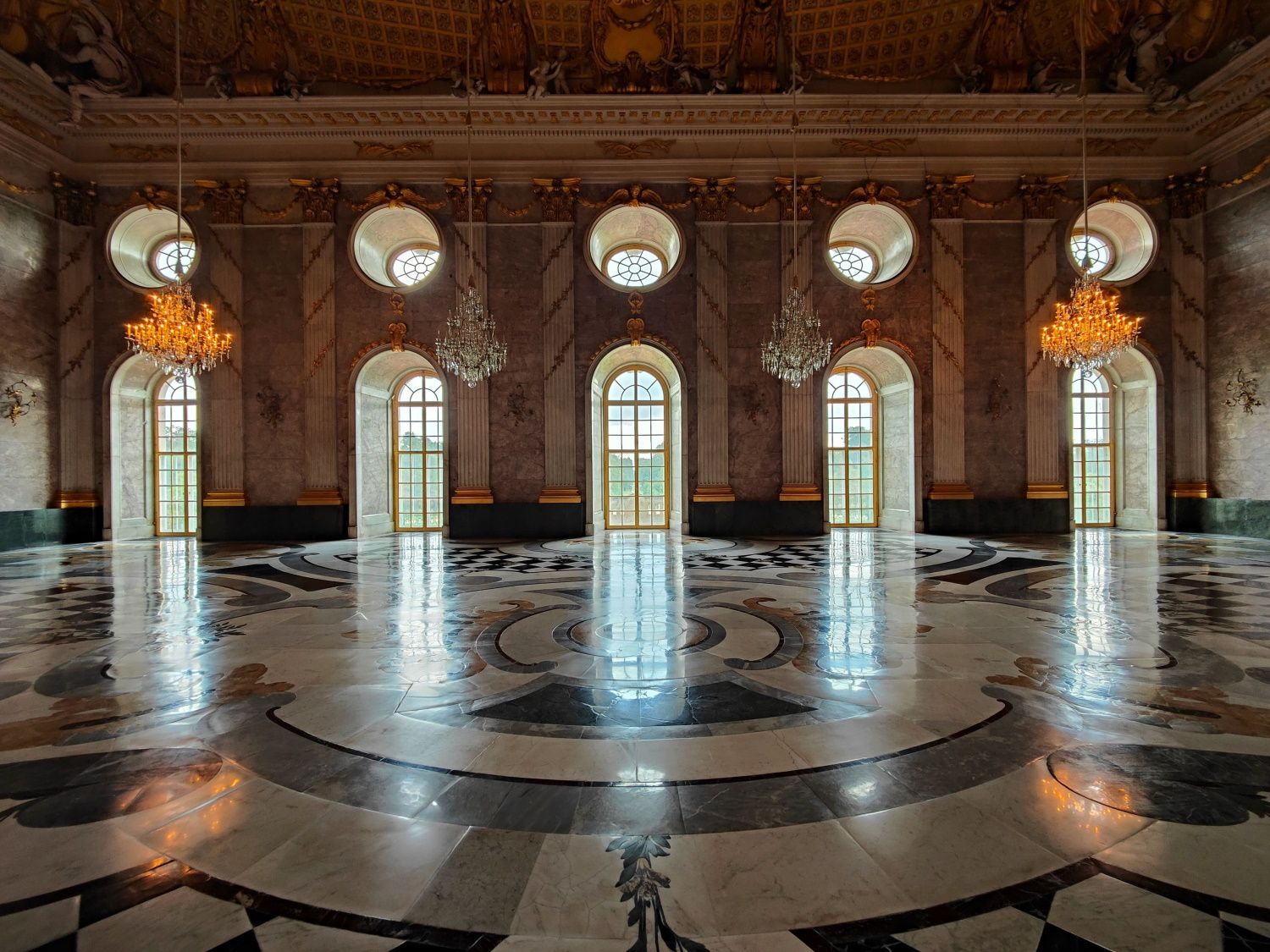 Ornate hall with marble floor, chandeliers, arched windows, and reflective surfaces.