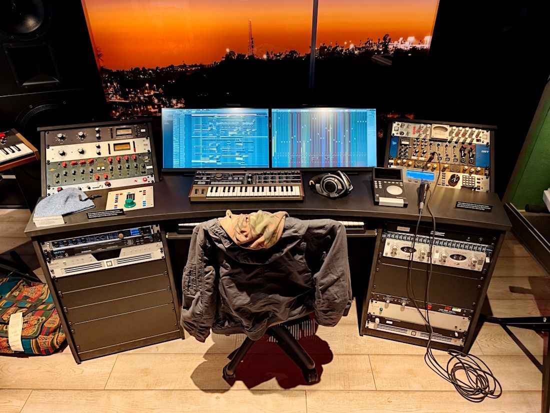 Dual-monitor audio editing station at sunset, with racks of gear and a central chair facing the desk
