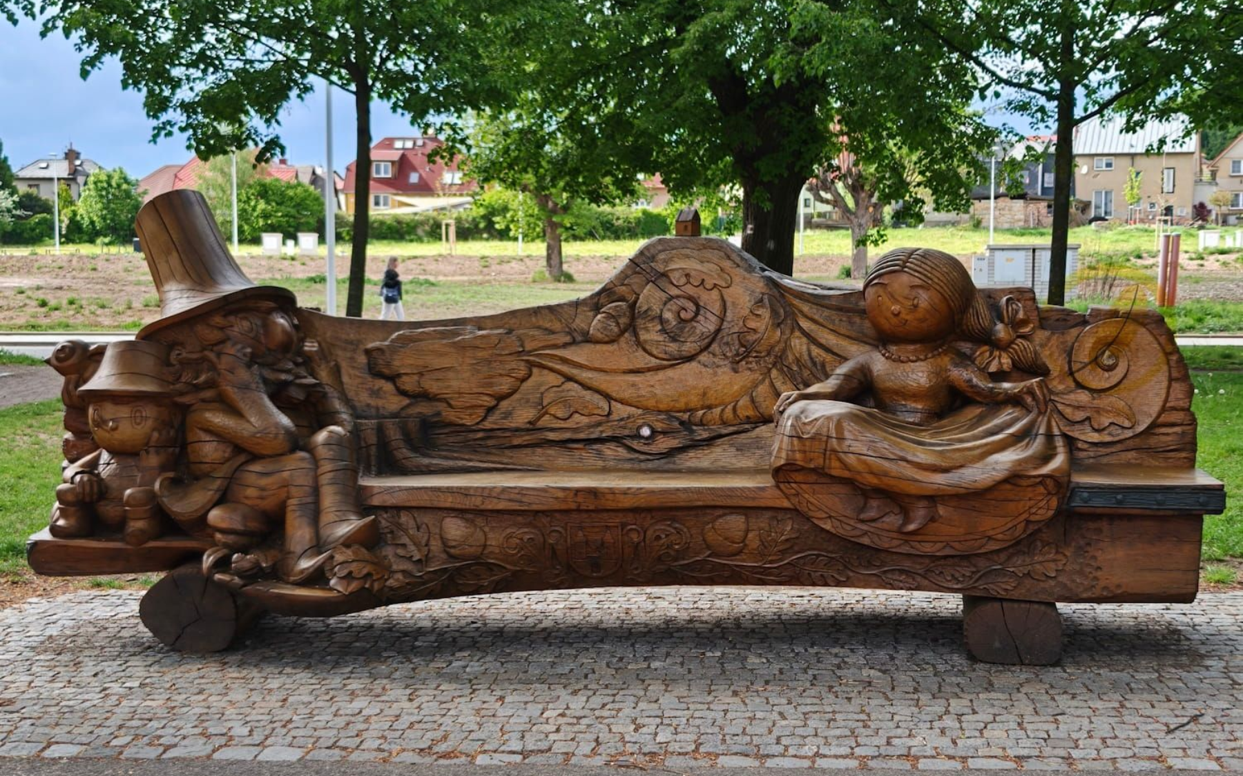 Bank in Jicín Fürchtenix Rust-colored sculpture of a reclining figure on a bench in a park, with trees and a walkway behind.