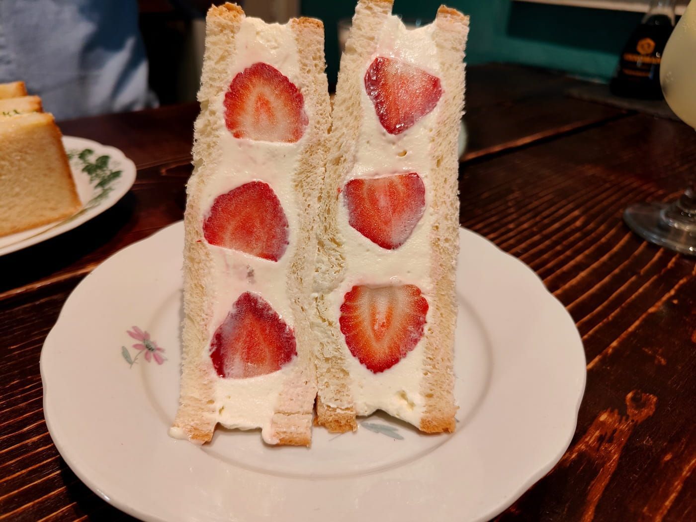Strawberry sandwich slices on a white plate on a wooden table, with another dessert in the background