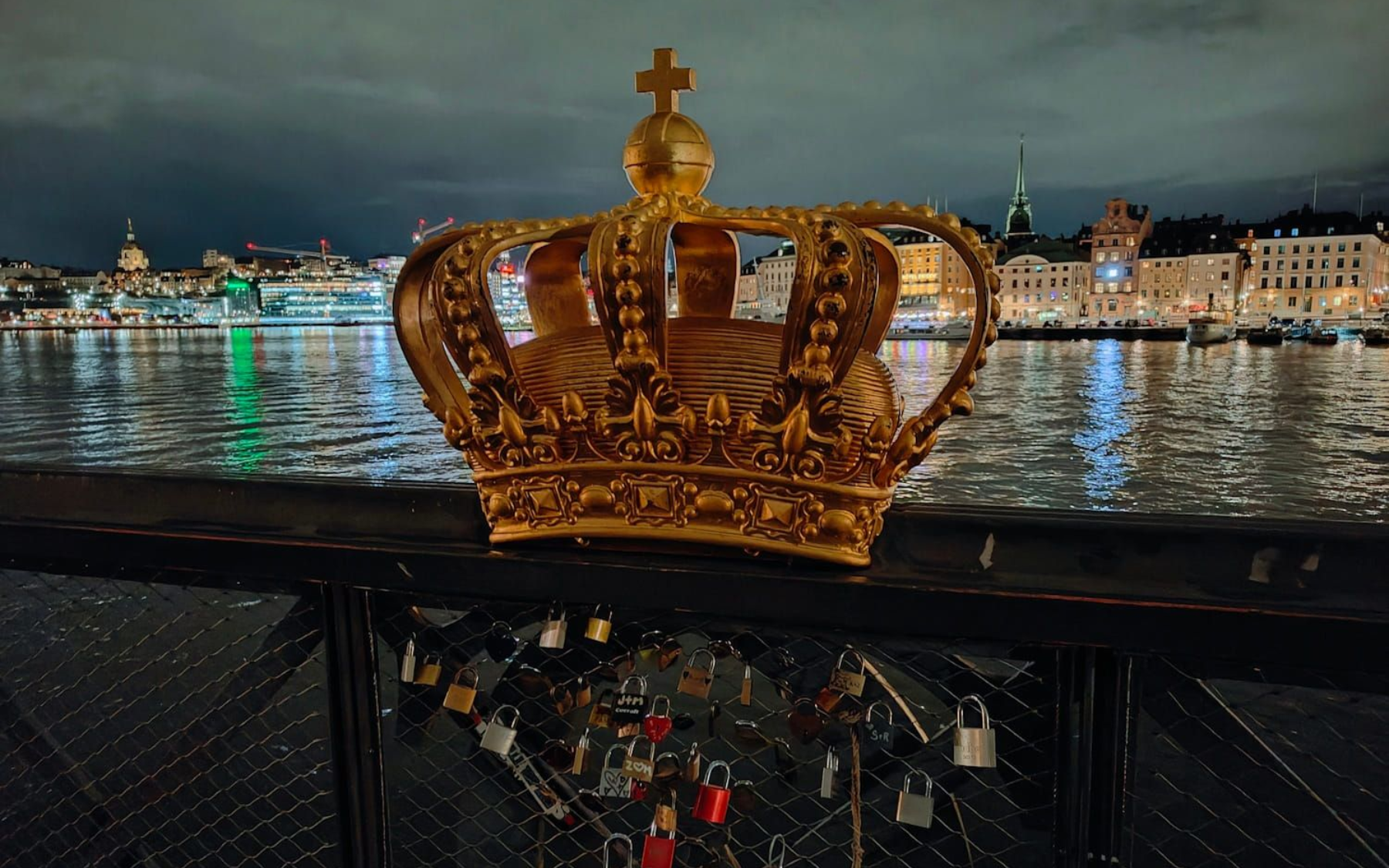 Eisenbrücke Skeppsholmsbron Golden crown on a riverfront railing at night, with city lights reflected on the water.
