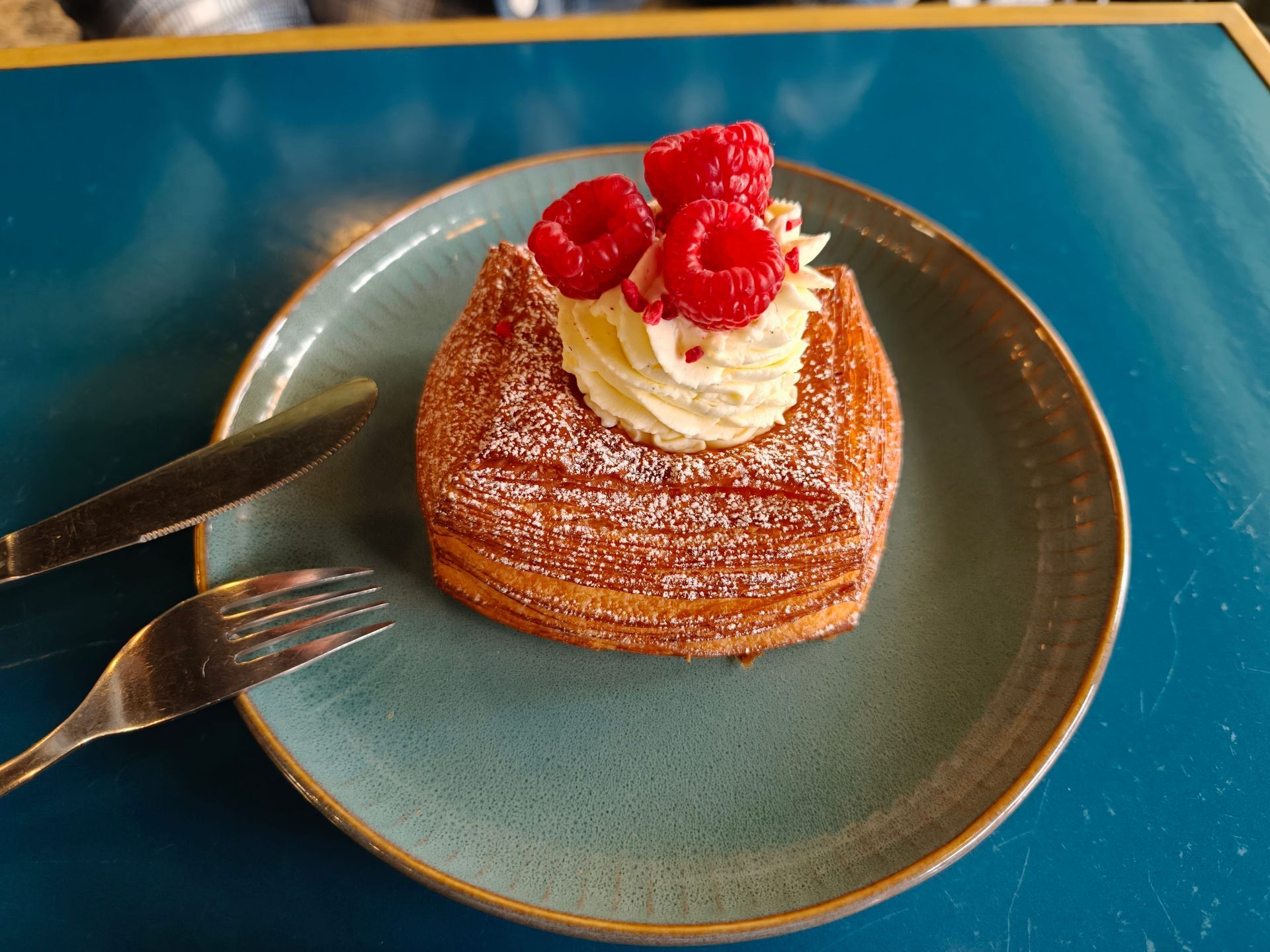 Pastry topped with whipped cream and raspberries on a blue plate with a fork and knife