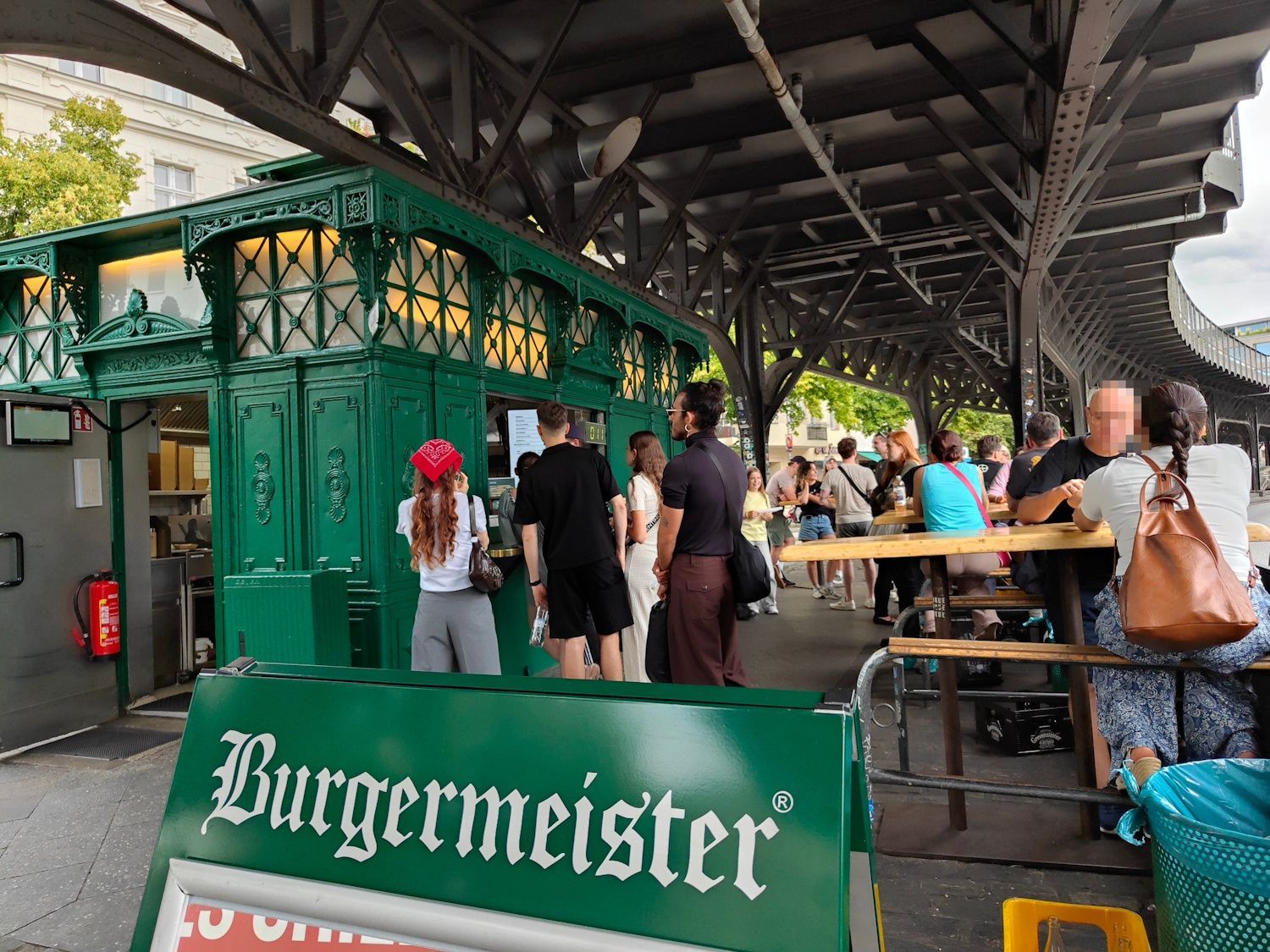 Burgermeister Berlin Covered outdoor beer garden with people seated at picnic tables under a green “Burgmeister” counter
