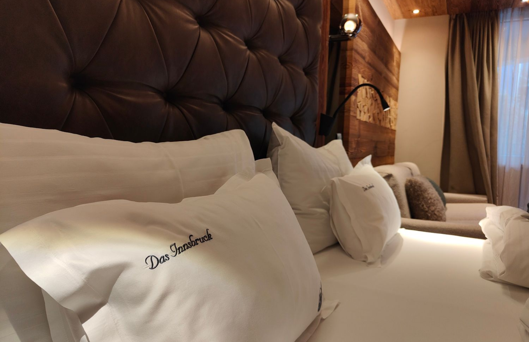 Das Innsbruck Cozy hotel bed with white linens, pillows, and a tufted leather headboard. Warm lighting and wood accents.