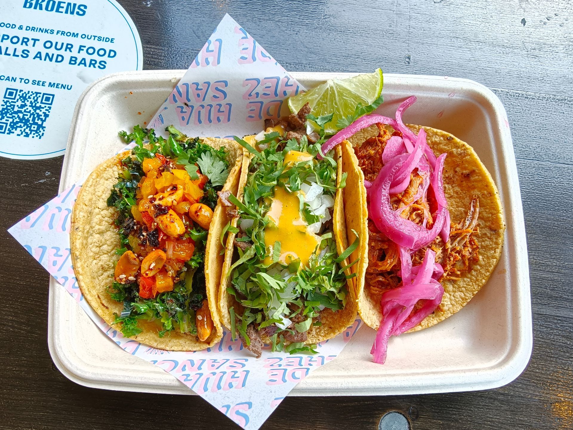 Three tacos on a takeout tray: orange, green, and pink fillings on corn tortillas.