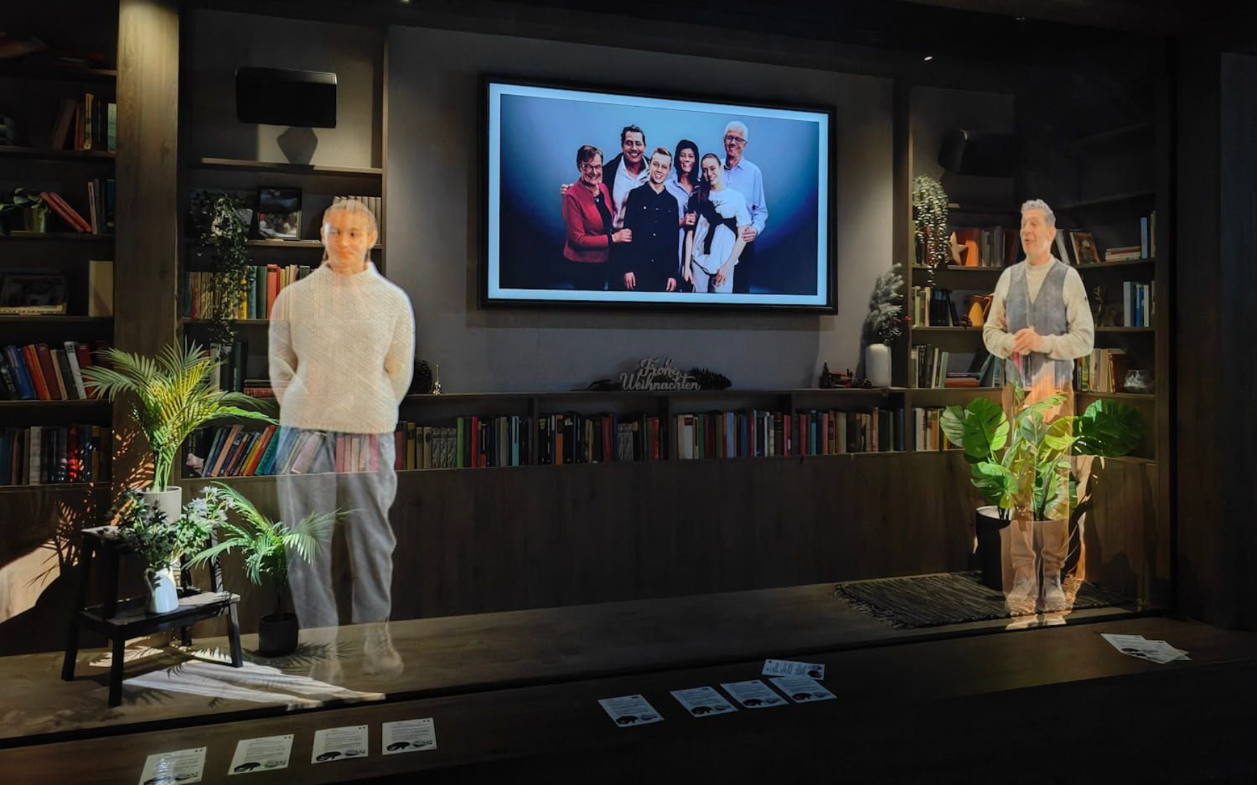 Experience Tirol Two figures stand on a stage, with a large screen displaying a family photo. Dark room with bookshelves.