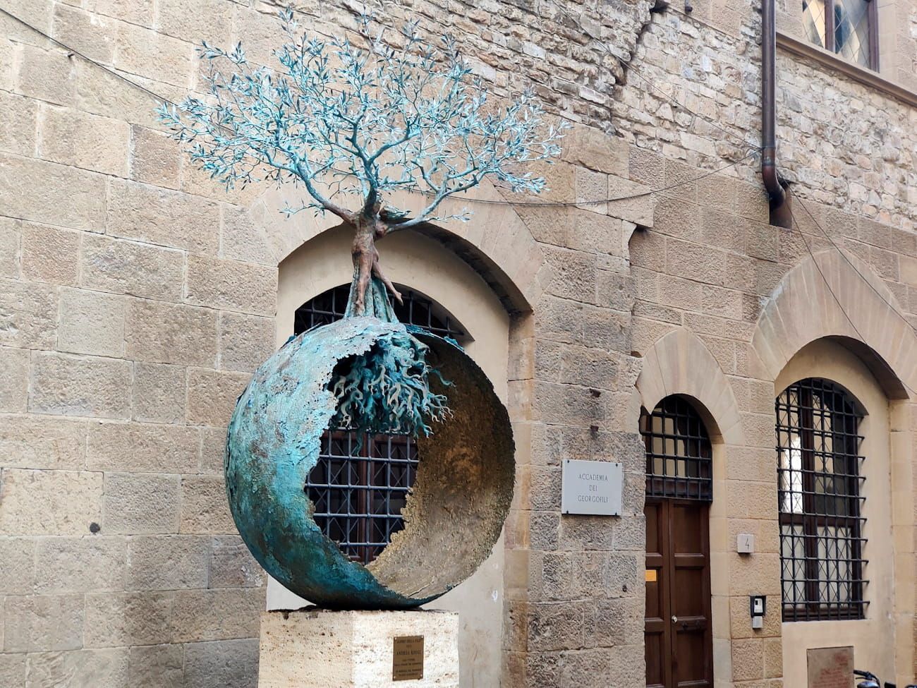 Baum des Friedens A bronze sculpture of a tree growing out of a hollow sphere, set against a stone building with arched doorways.