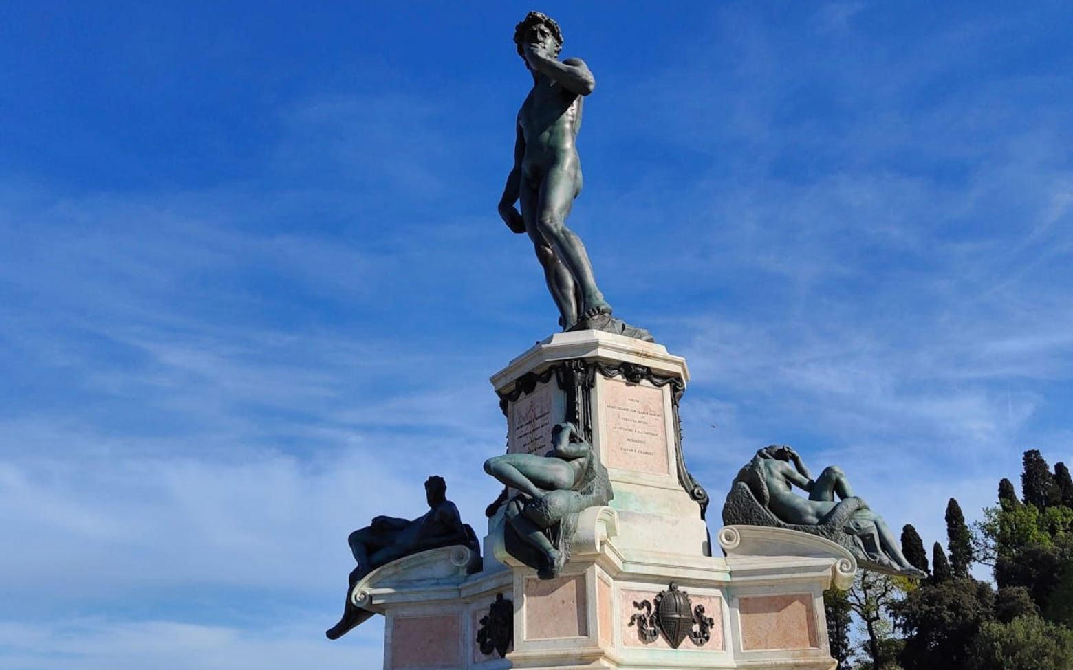 Bronze statue of David Bronze statue of David atop a stone pedestal with reclining figures, set against a bright blue sky.