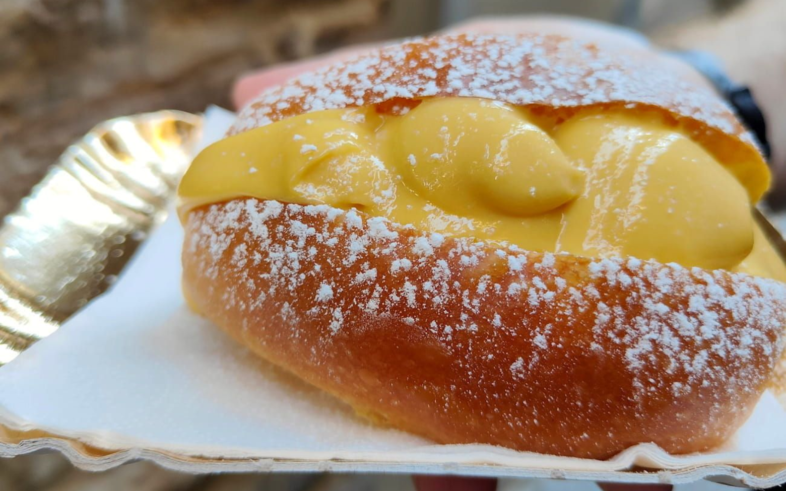 Minuto A soft, golden-brown brioche bun filled with smooth yellow cream and dusted with powdered sugar on a paper tray.