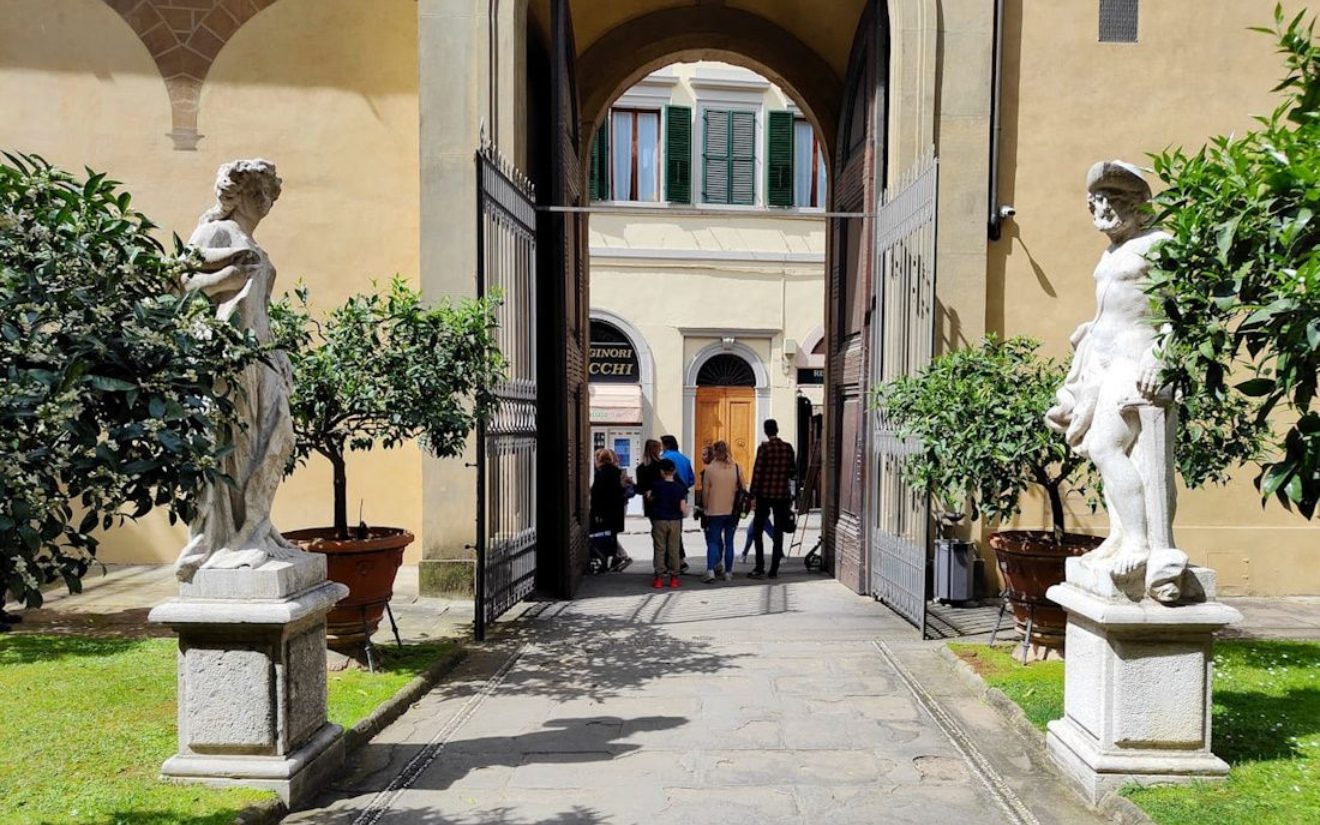 Florenz Medici Garten A stone walkway leading through a gate flanked by two statues and potted plants toward a courtyard building.