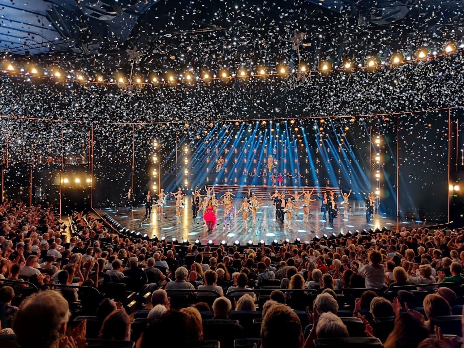 Concert stage with performers under blue lights and confetti, seen from a packed audience under string lights