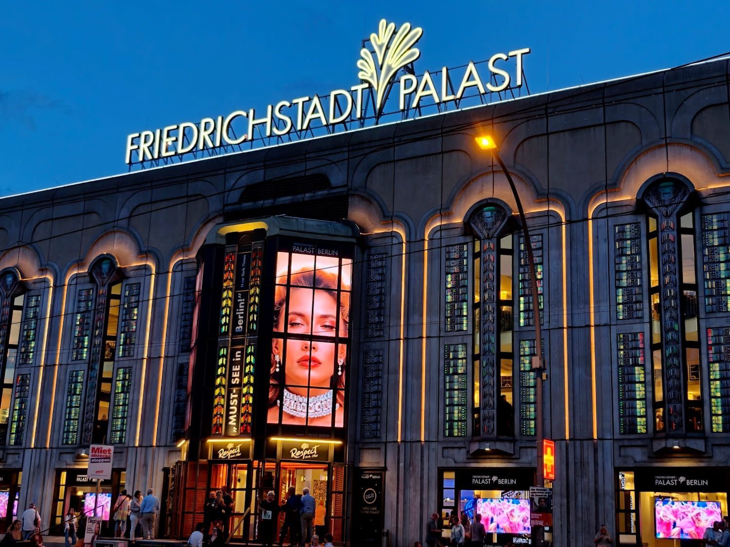 Friedrichstadt Palst Berlin Friedrichstadt-Palast theater façade at dusk, lit in blue and gold with tall illuminated windows and a digital face display