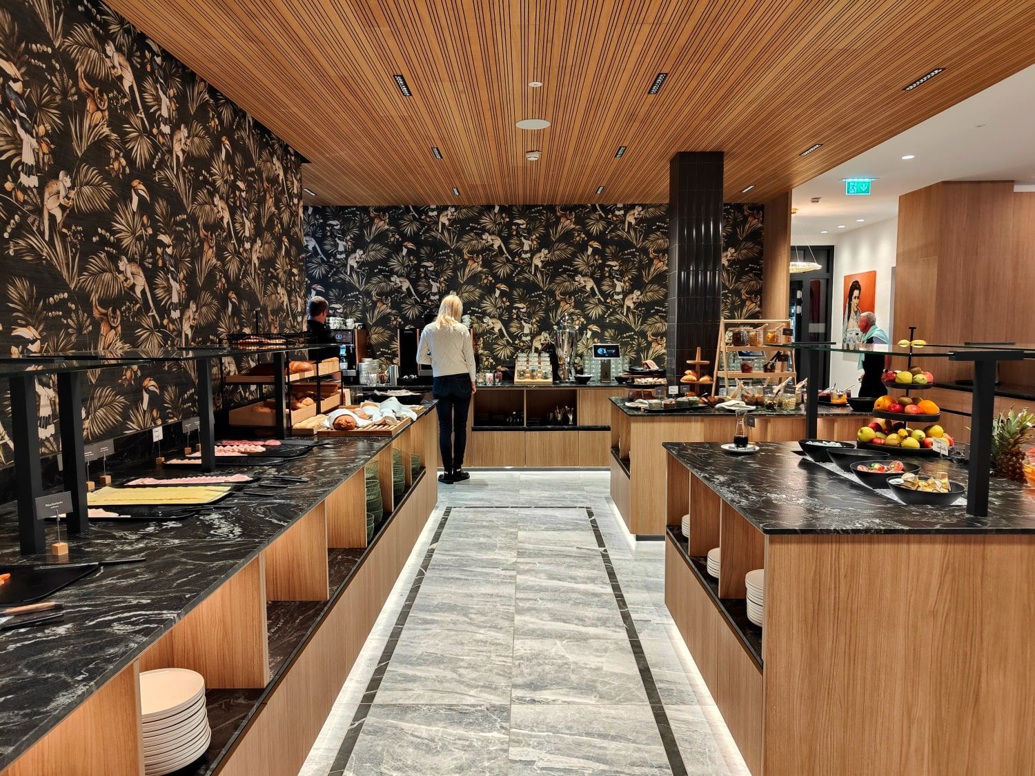 Buffet area with food displays. People stand near tables with black countertops, wood accents, and patterned wall.