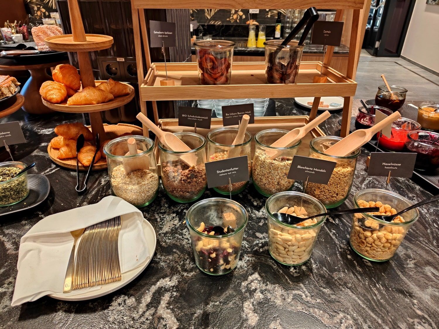 Breakfast buffet with various cereals, toppings, pastries, and jams, on a dark countertop.