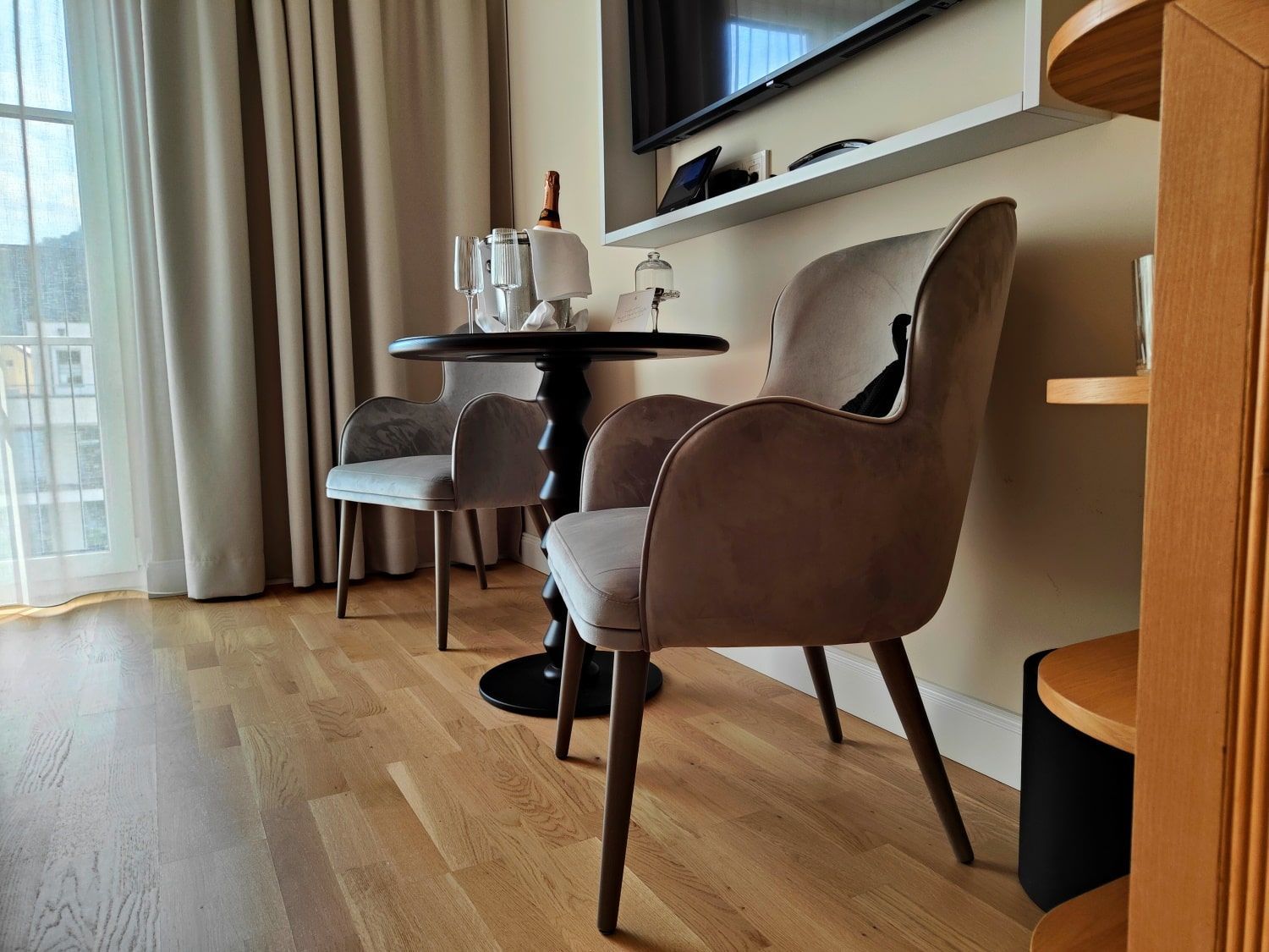 Hotel room interior: table, chairs, view to left through window. Light colored wood floor.