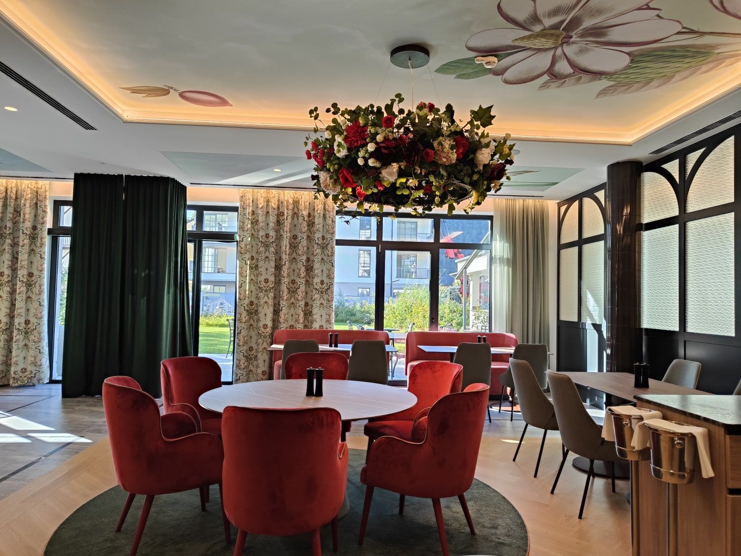 Elegant dining room with red chairs, round table, floral chandelier, patterned curtains, and garden view.