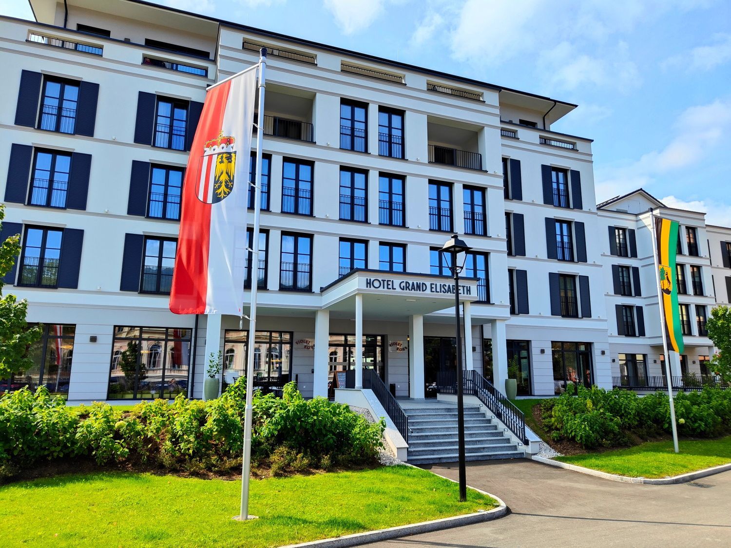 Grand Elisabeth Hotel Hotel exterior with flags, white building with blue shutters, and a green lawn.