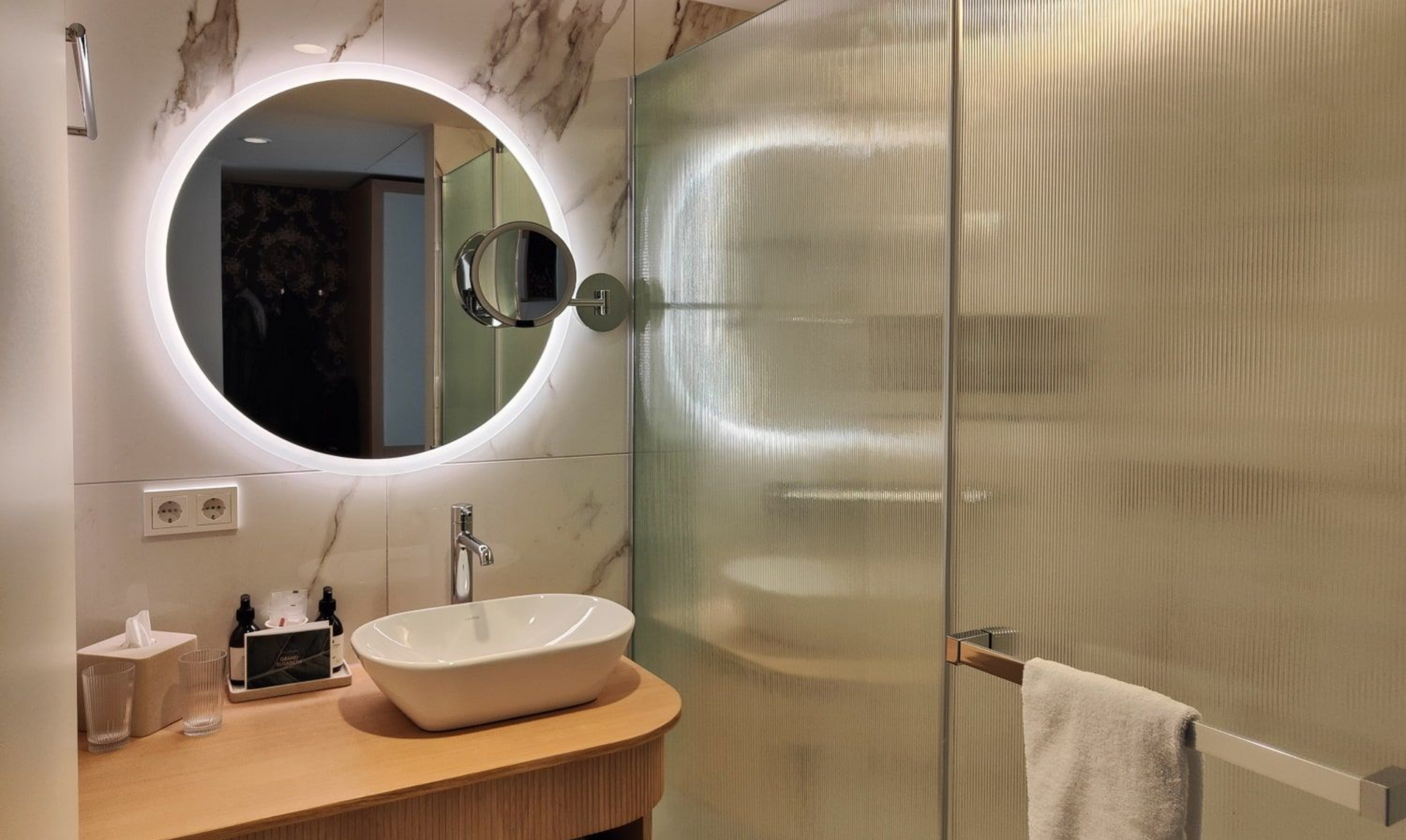 Grand Elisabeth Hotel Bad Modern bathroom with lit round mirror, vessel sink, glass shower, and marble walls.