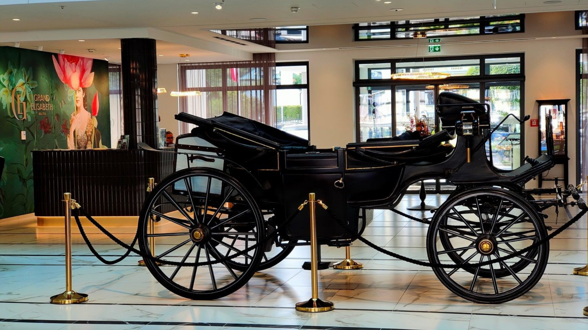 Grand Elisabeth Hotel Kutsche Black horse-drawn carriage displayed in a hotel lobby, behind a golden rope barrier.