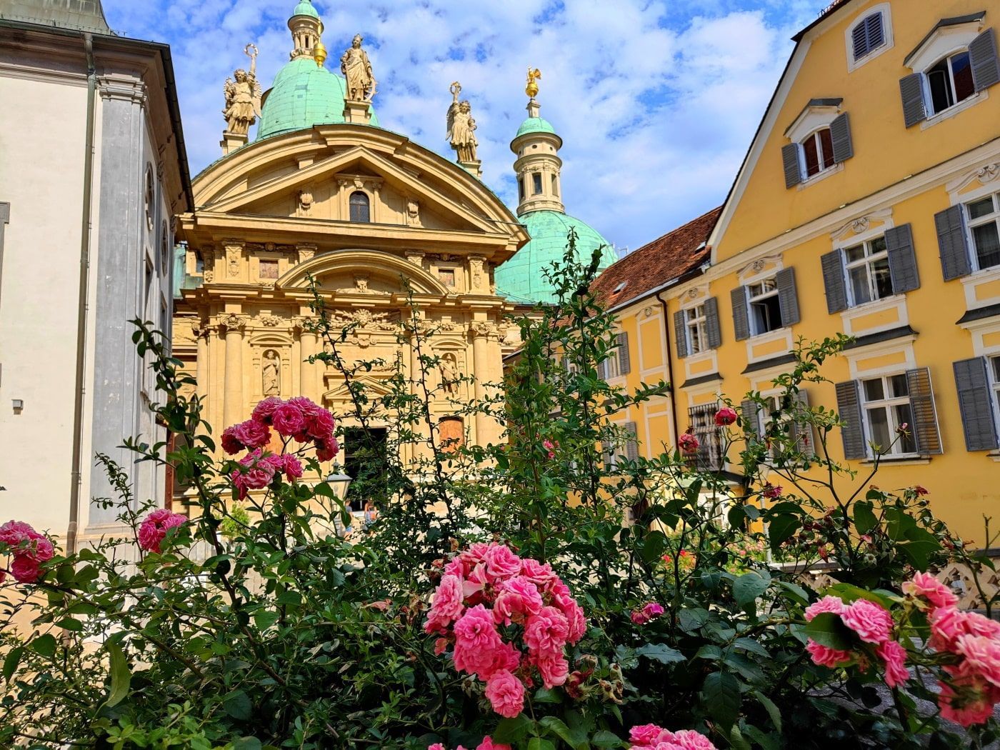 Graz Pink roses in front of an ornate yellow building with green domes and a yellow building to the right under a blue sky.