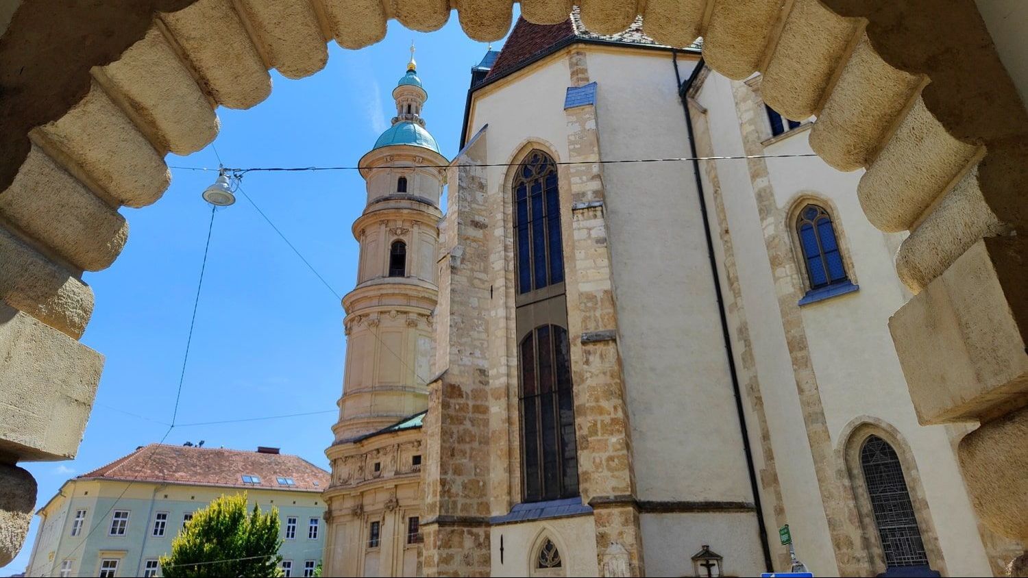 Graz View through an arched doorway of a tall church with a green-topped tower against a blue sky.