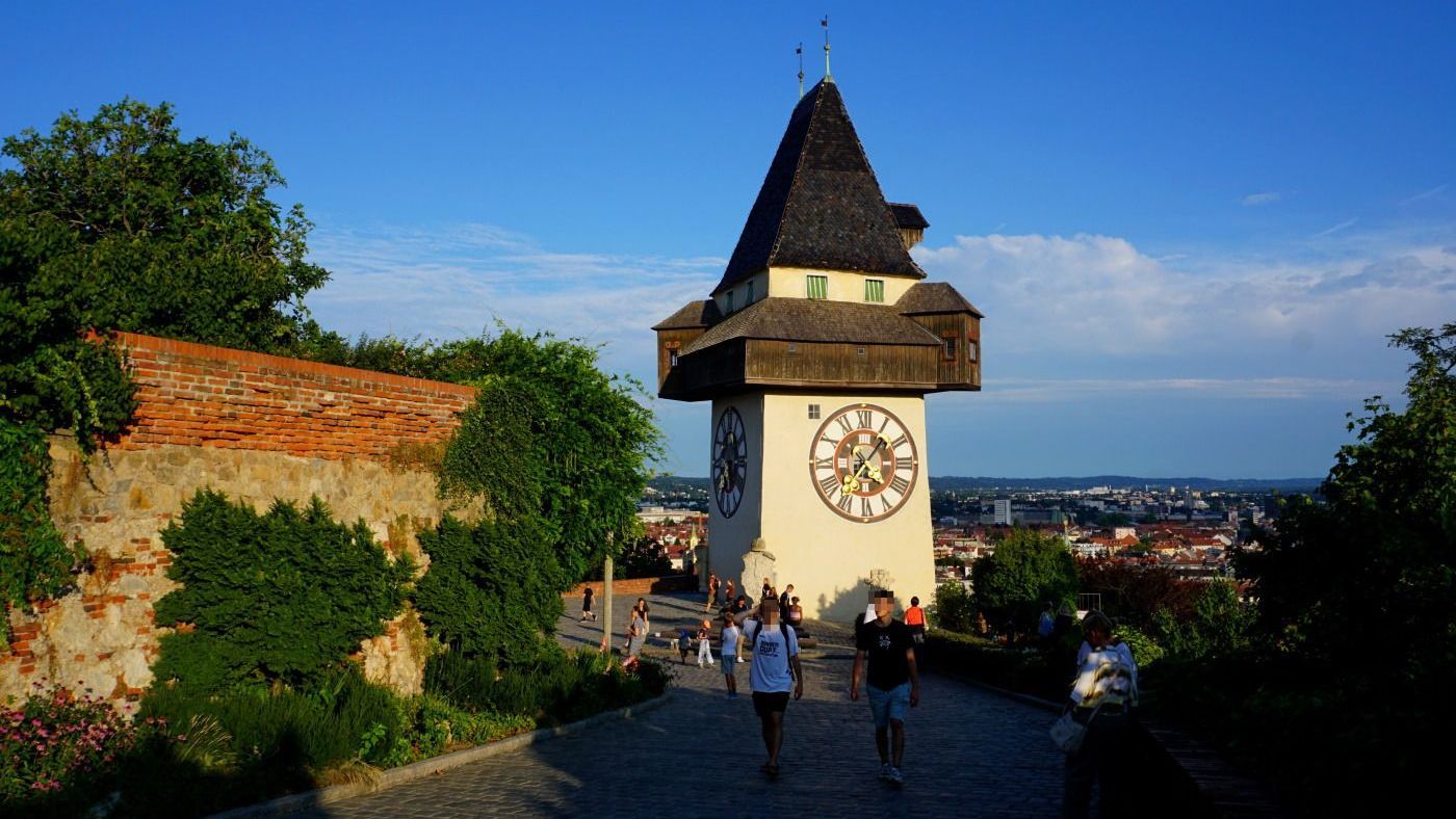 Graz Clock tower in Graz, Austria, with a prominent clock face, overlooking the city on a sunny day. People walk on the pathway.