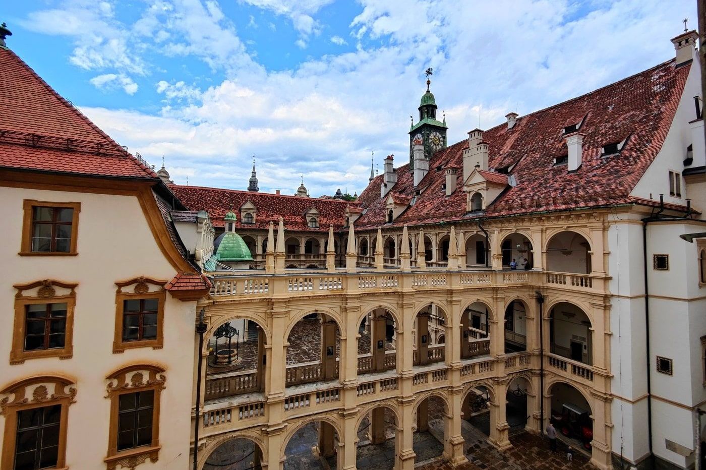 Graz Courtyard of Eggenberg Palace in Graz, Austria, with arched walkways, red-tiled roofs, and a blue sky with scattered clouds.
