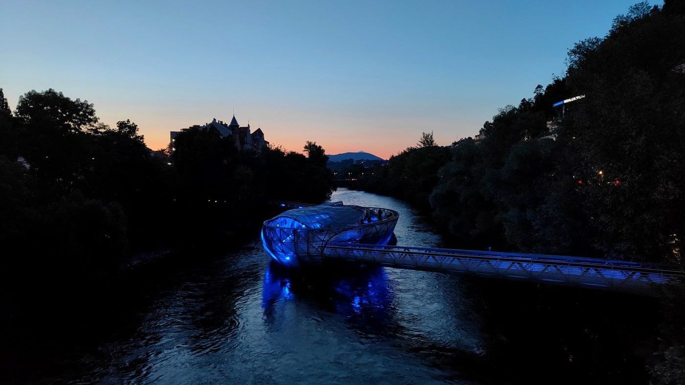 Graz A river with a blue-lit structure flows through a dark forest at dusk. Distant buildings are silhouetted in the orange and blue sky.