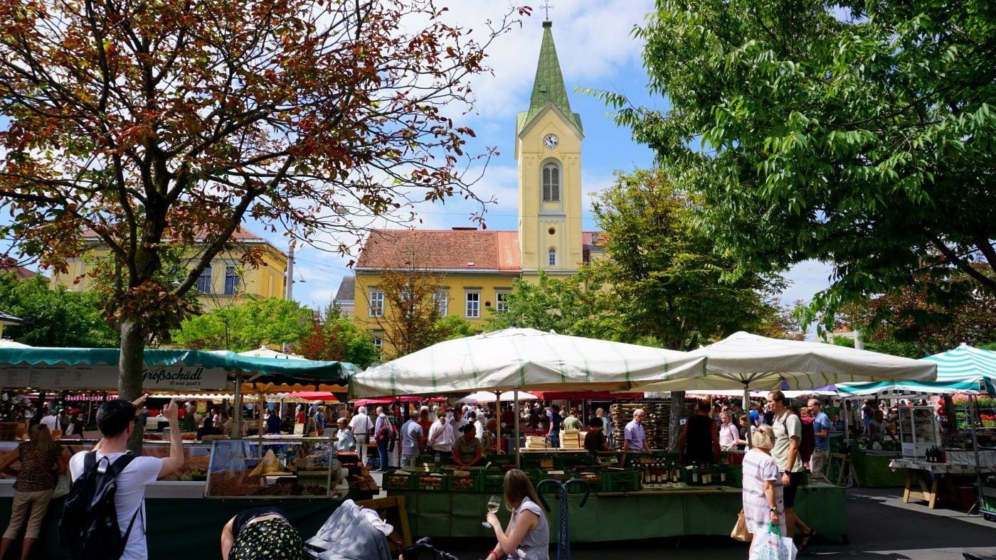 Marketplace bustling with people and vendors, with a church spire rising in the background under a partly cloudy sky.