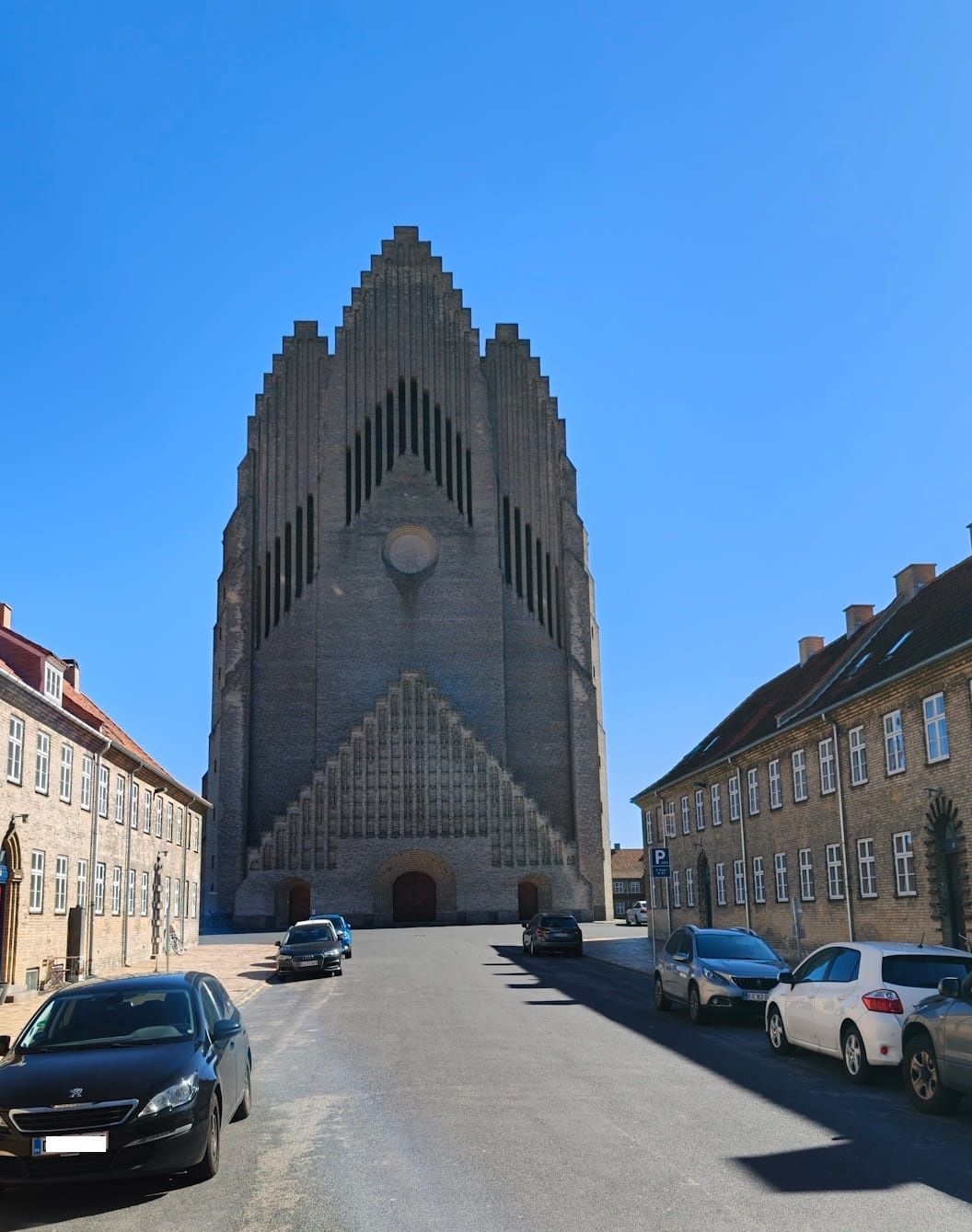 A tall Gothic cathedral viewed from a street lined with parked cars under a clear blue sky.