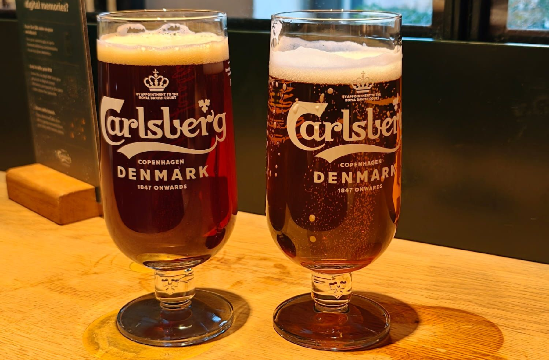Home of Carlsberg Bier Two Carlsberg beer glasses on a pub table, one full and one partially filled, with foam on top. Home of Carlsberg