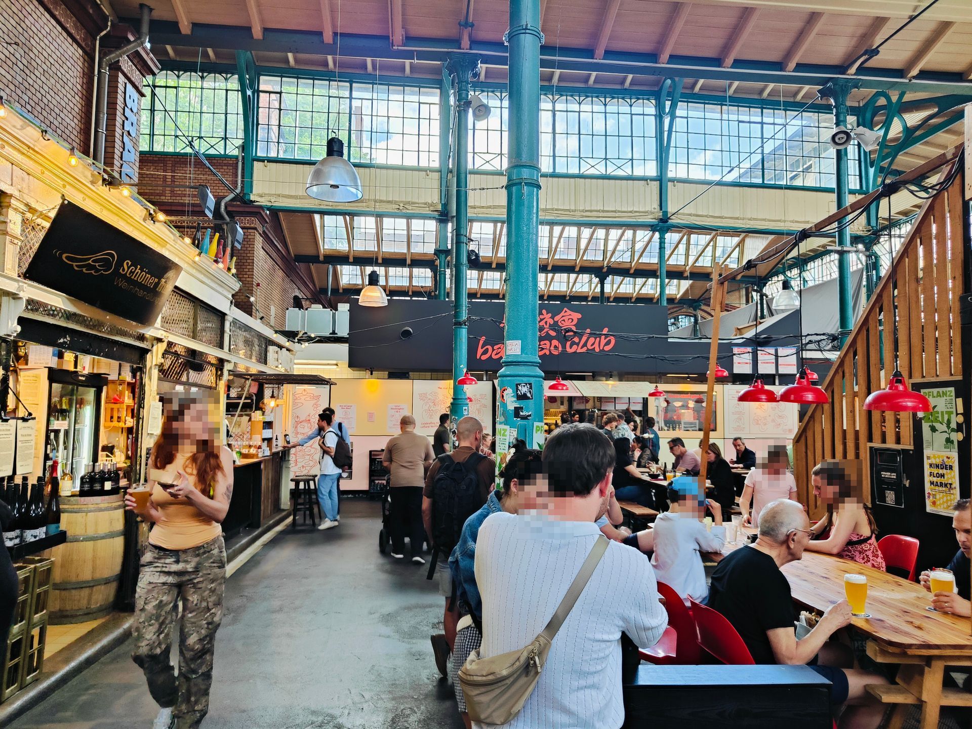 Bustling indoor food hall with diners at tables under bright skylights and hanging red lamps