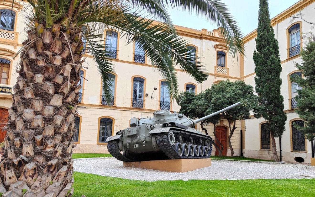 Valencia Heeresmuseum Tank statue on a platform in front of a building with windows, palm tree in foreground.