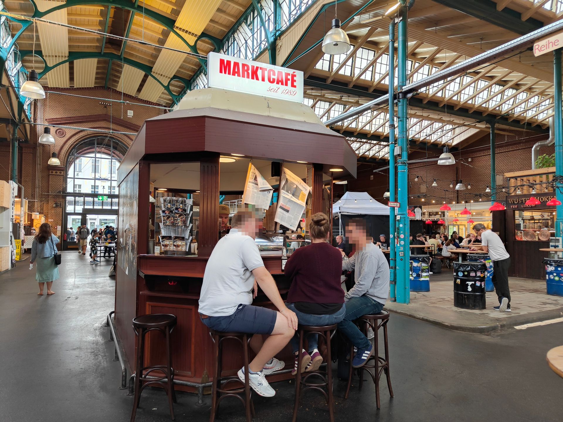 People seated at a small café stand in a large indoor market hall with a glass roof.