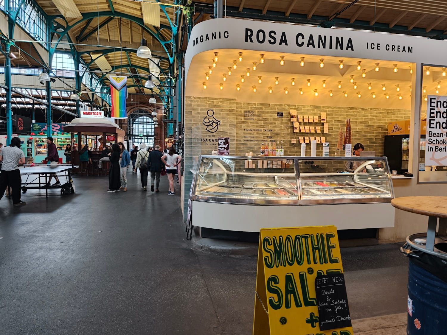 Market stall named Rosa Canina in an indoor hall, with people walking and a yellow sale sign in front
