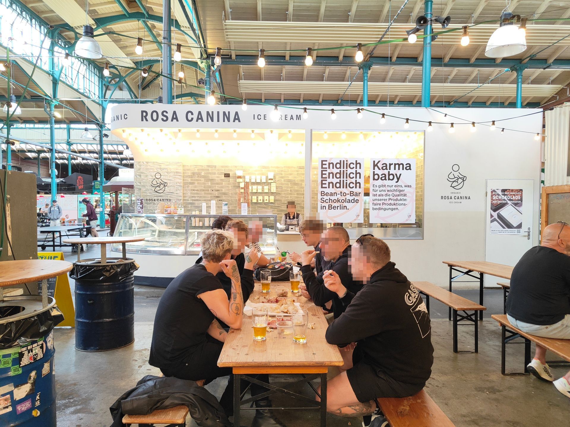 People seated at a long table in a bright indoor market café with hanging lights and white walls