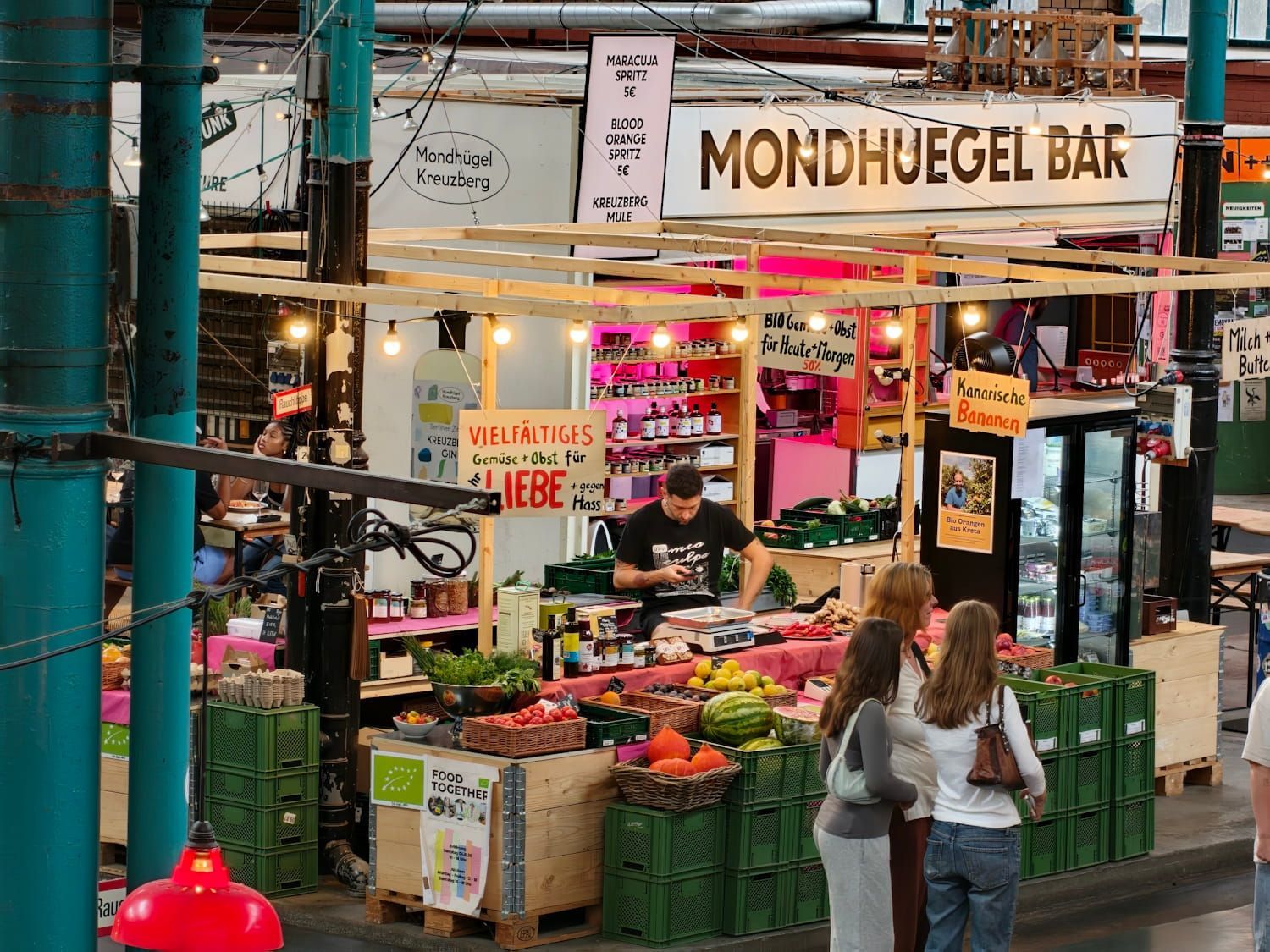Open-air market stall with fruit crates, string lights, and two people browsing under a “MONDVGEL BAR” sign