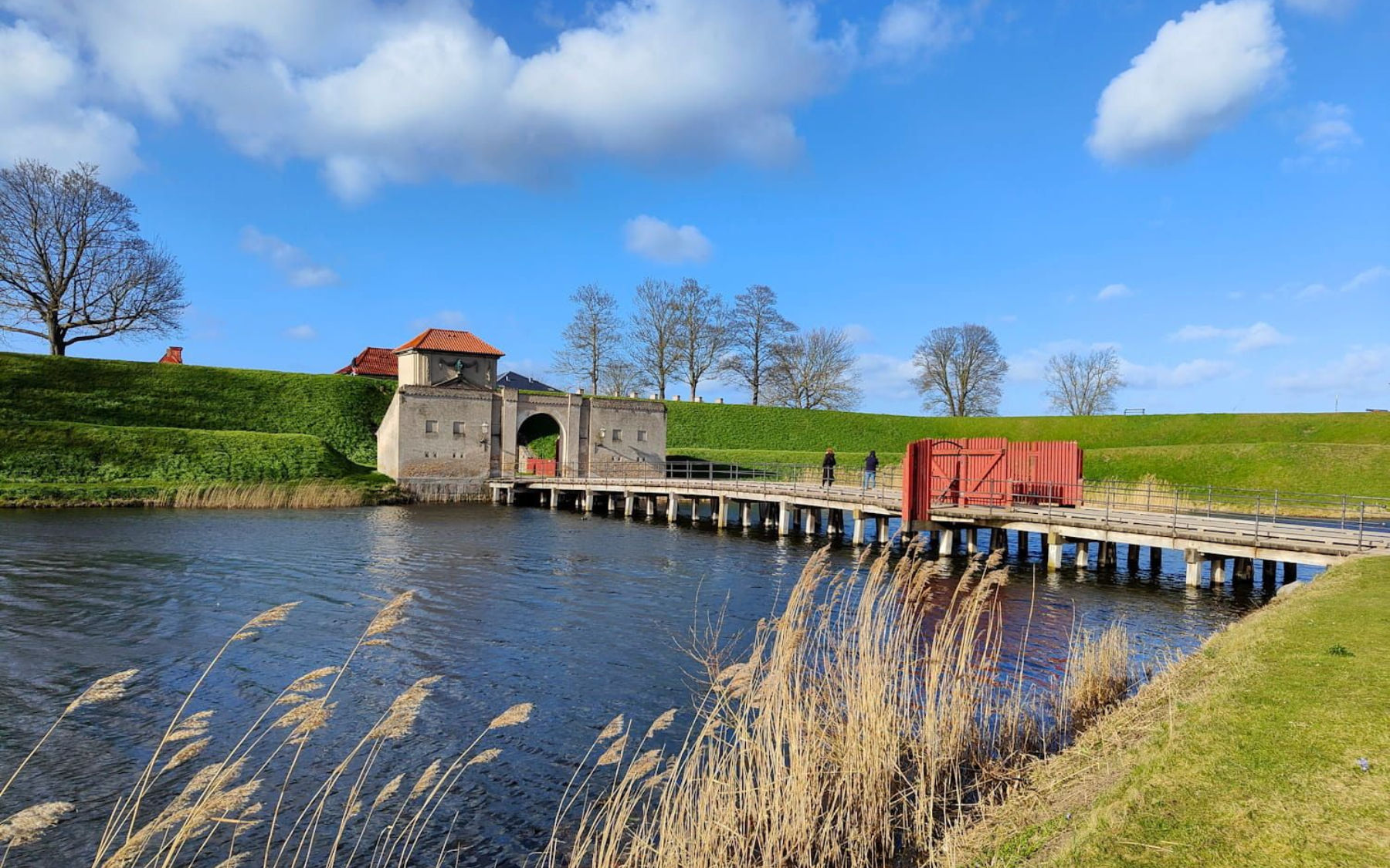 Kastell von Kopenhagen Stone bridge over a canal with reeds in front, a red sign, grassy banks, and a small tower under a blue sky. Kastell von Kopenhagen