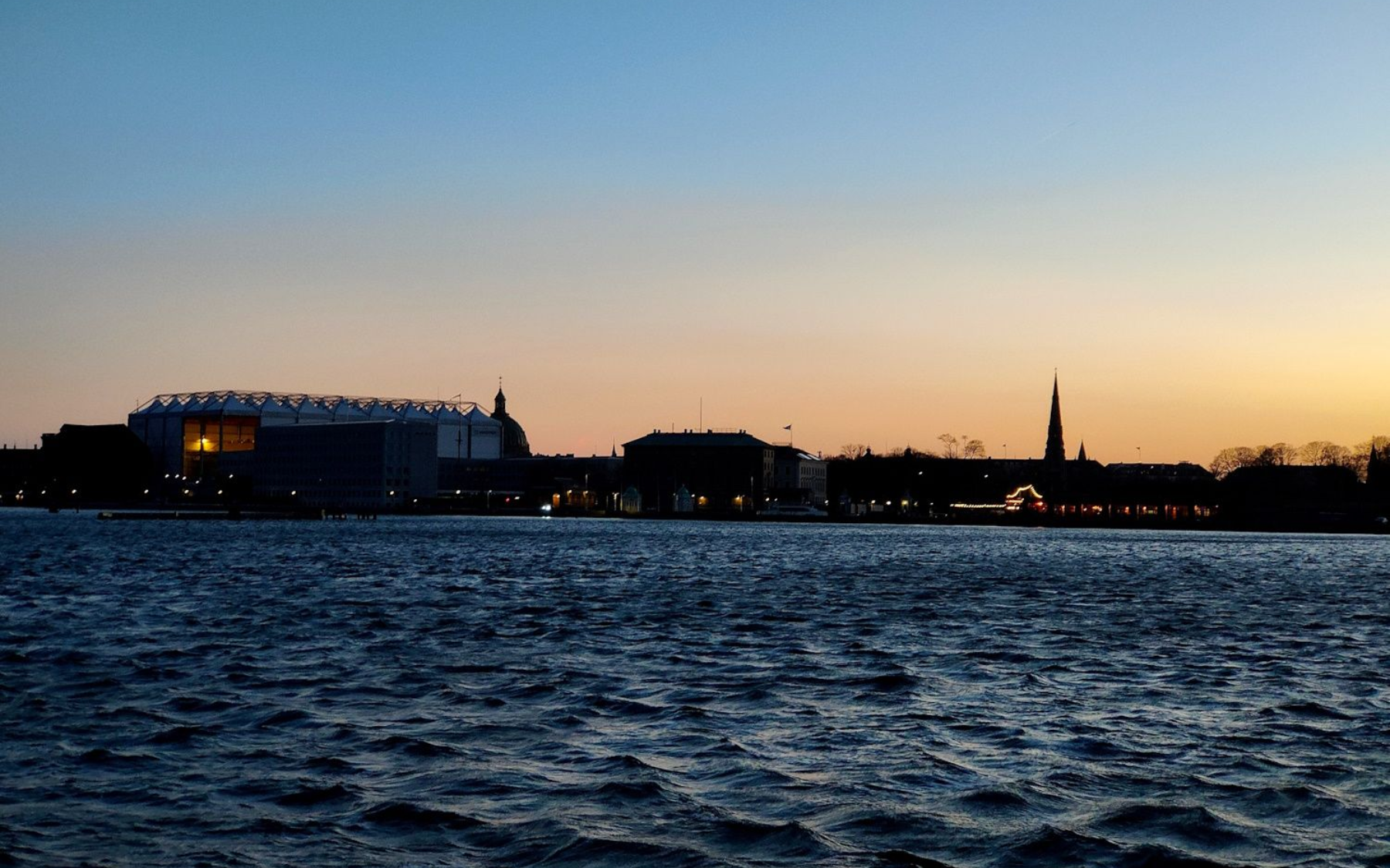 Kopenhagen Skyline Dusk over a choppy river with city skyline silhouetted against a blue-orange sky and a church spire.
