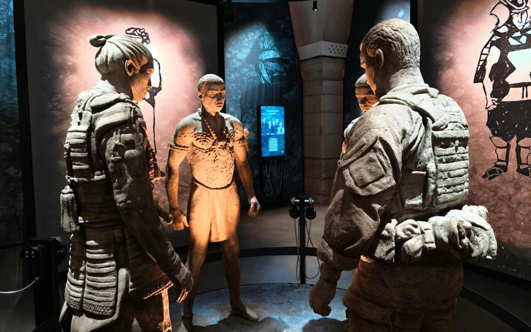 Museum Three people in futuristic armor stand in a sci-fi exhibit room with glowing displays and stone-like walls