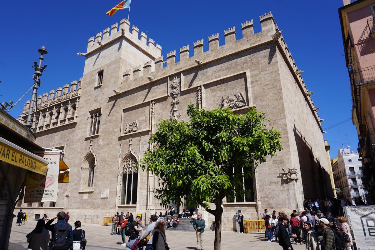 A tall stone building with battlements, a tower, and a flag, people gather in a square.