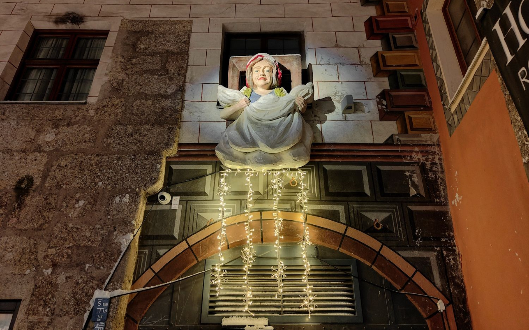 Märchengasse Statue of angel above an arched doorway, lit with fairy lights, on an old building at night.