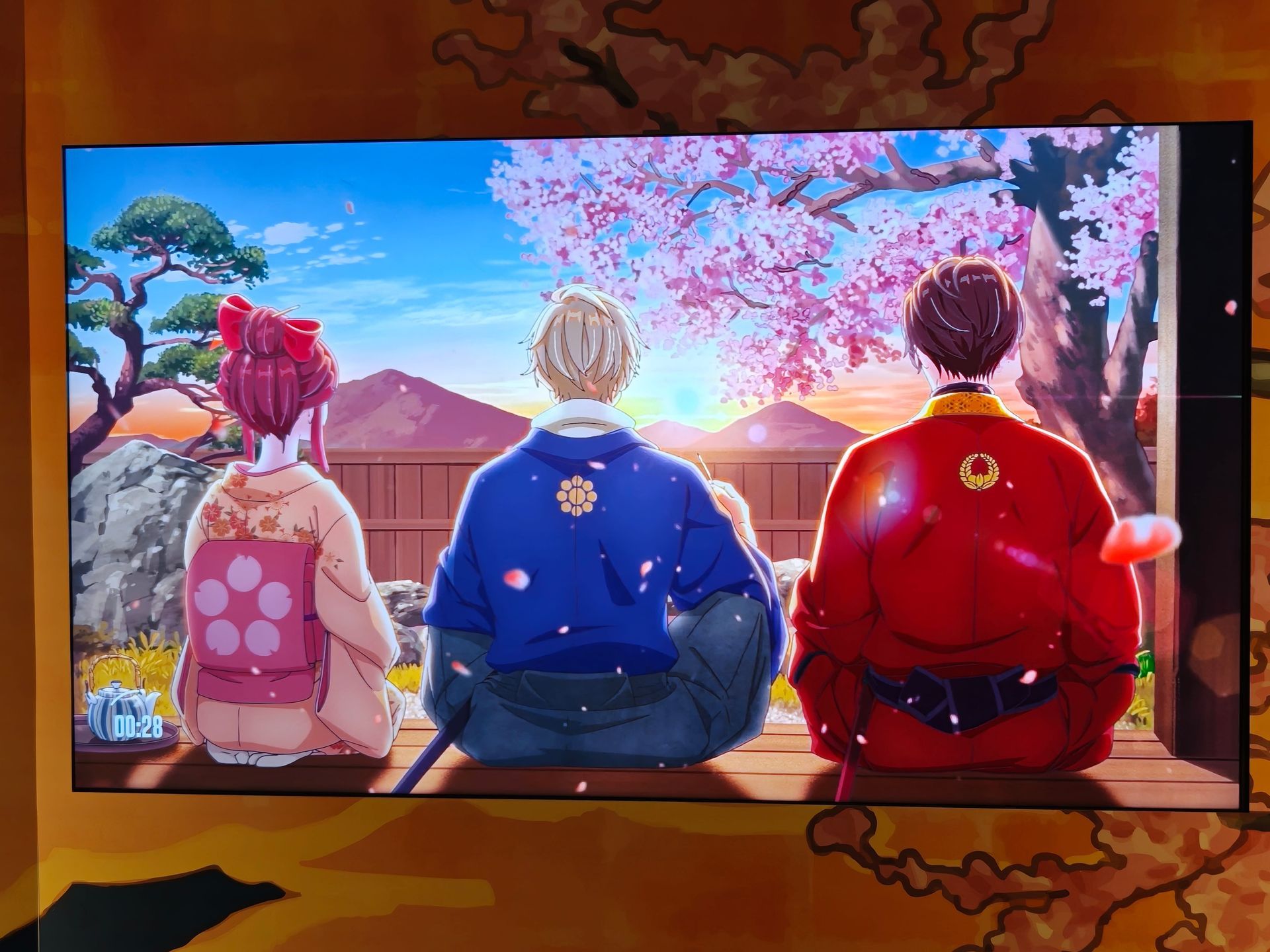 Three robed figures sit on a hillside, facing a sunrise with cherry blossoms and a distant mountain.