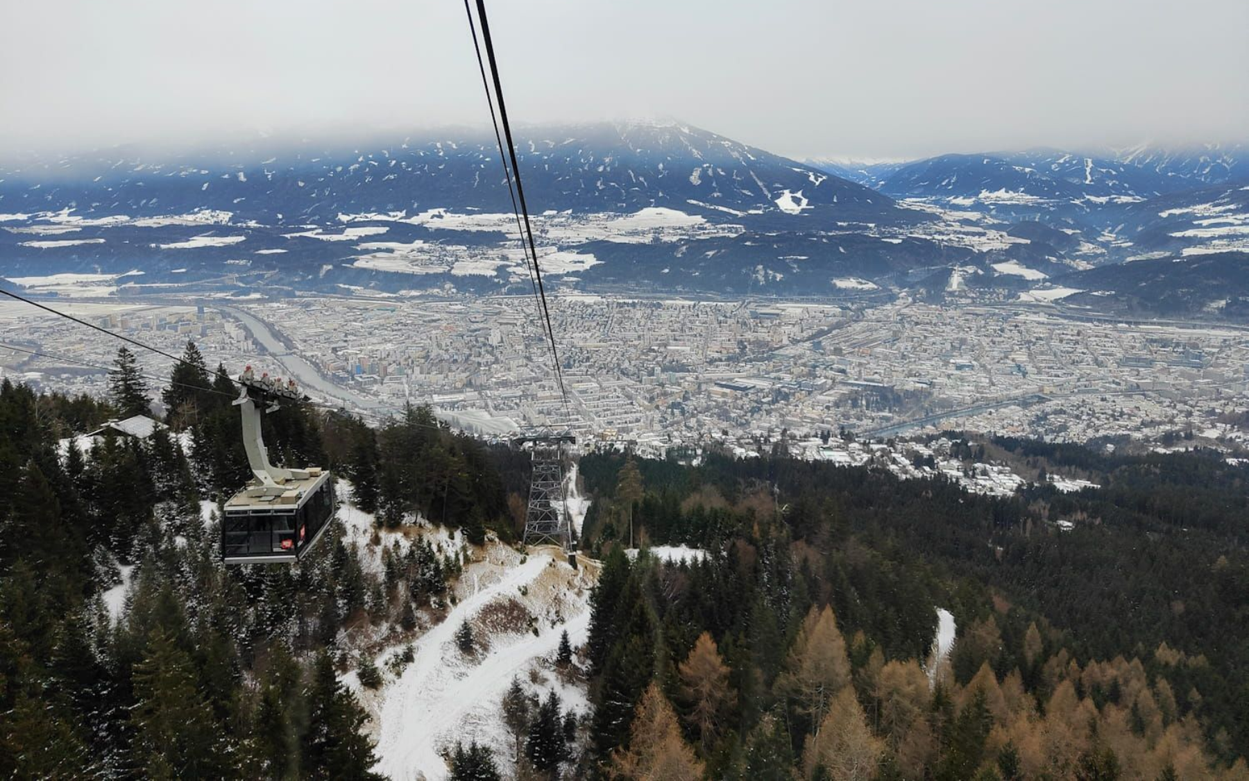 Nordkette Innsbruck Cable car over snowy town and forest, cloudy sky.