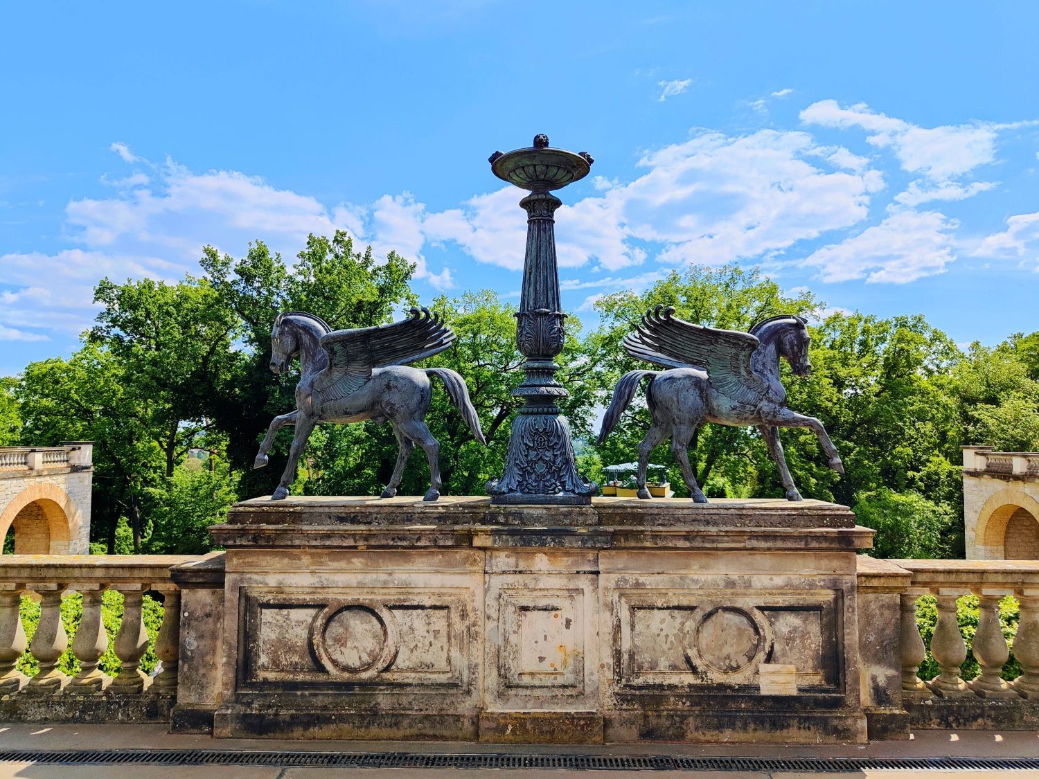 Bronze horse sculptures flank a fountain on a stone terrace, set against a backdrop of trees and blue sky.