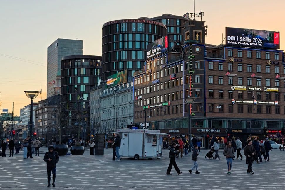 Crowded city square at sunset with pedestrians, modern buildings, and a white truck in the foreground