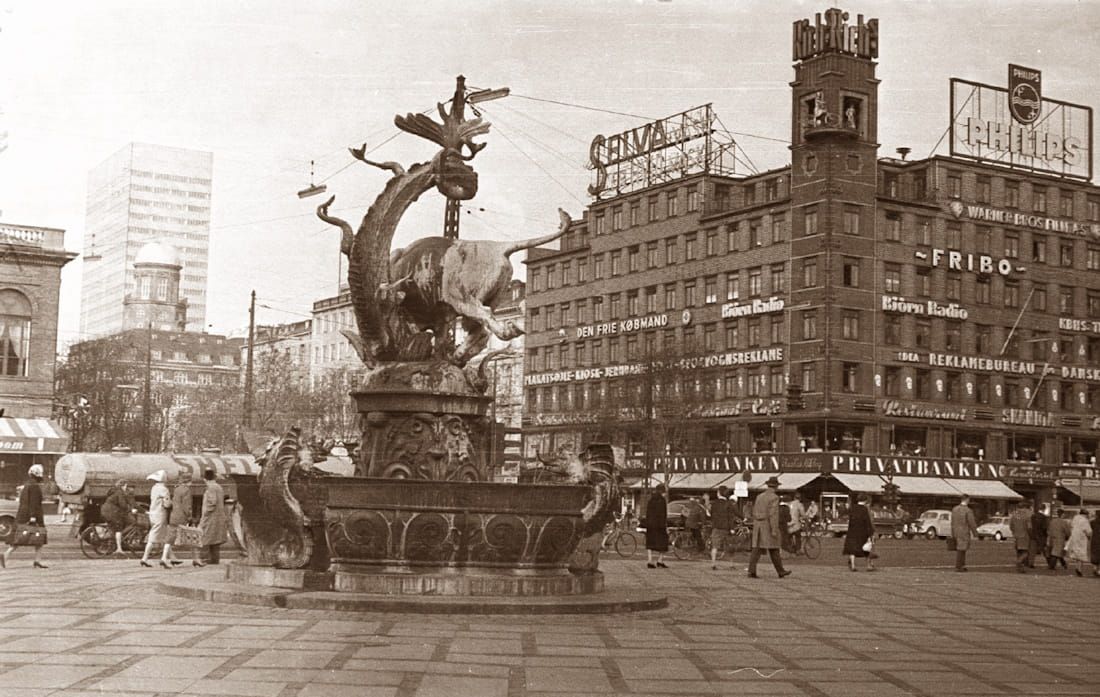 Sepia city square with a fountain sculpture and busy pedestrians beside old buildings