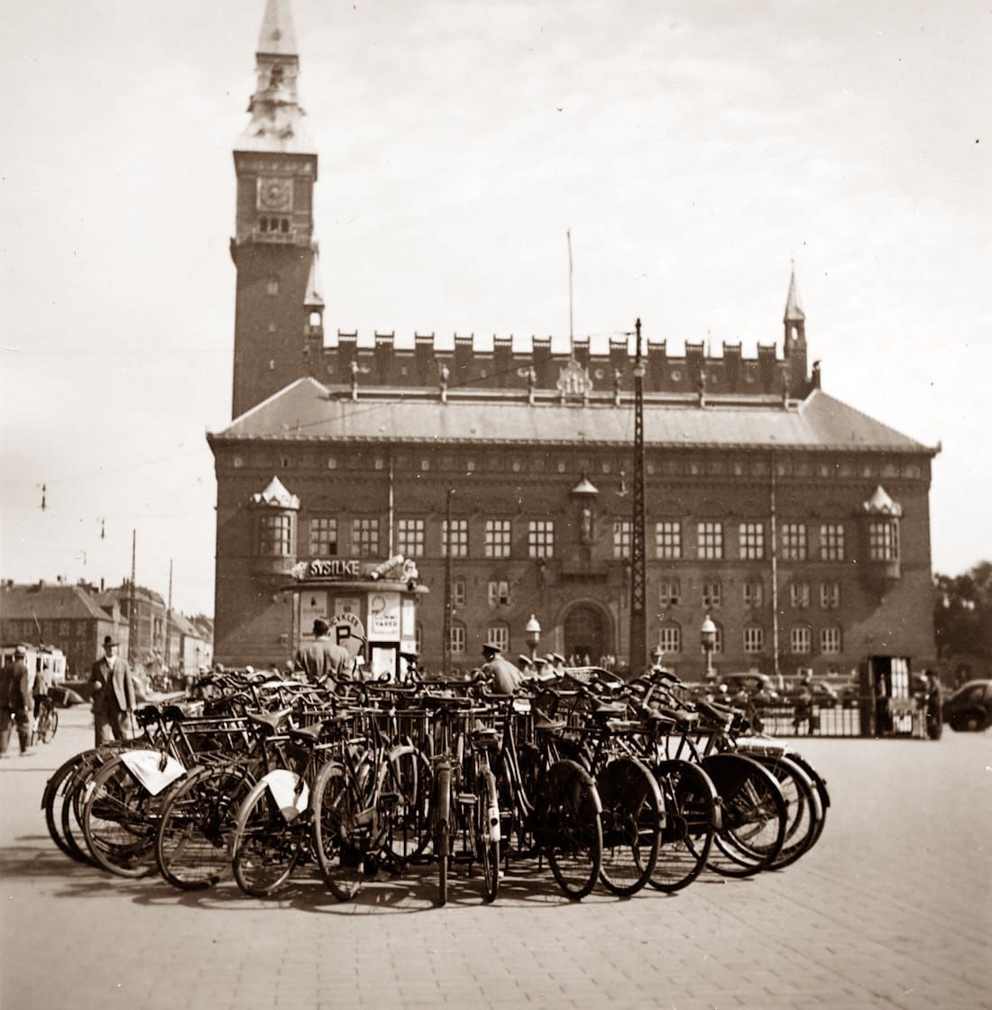 Crowded bicycle parking in front of a historic brick building with a tall clock tower